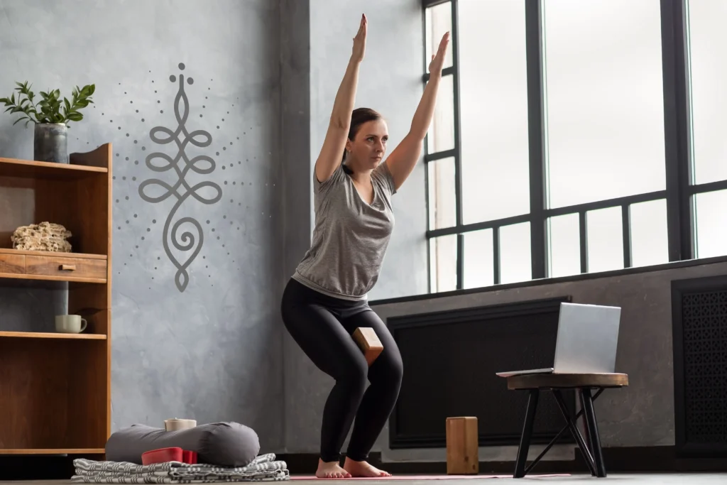 Femme caucasienne, dans la quarantaine, qui fait du Yoga (posture debout, Utkatasana dite la posture féroce) via des cours en ligne. Elle utilise un bloc de bois entre ses cuisses pour aider à contracter ses quadriceps.
