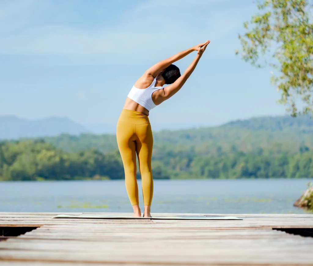 Femme de dos, en posture debout, en train d'étirer les muscles de son flanc gauche. Ses hanches sont décalées vers la gauche et ses bras, allongés vers le ciel, tendent vers la droite. Elle est située devant un plan d'eau avec vu sur les montagnes.